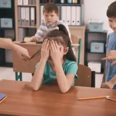 Dziewczynka zakrywa twarz, gdy inne dzieci dokuczają jej w szkole. Jedno ciągnie ją za włosy, inne wskazują palcem.