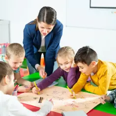 Nauczycielka z dziećmi rysuje mapę. Dzieci spędzają czas w przedszkolu, co może wiązać się z opłatą za przedszkole powyżej 5 godzin.