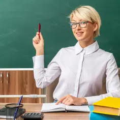 Nauczycielka z czerwoną kredą w dłoni, siedząca przy biurku, wyjaśnia, jak obliczyć pensum łączone.