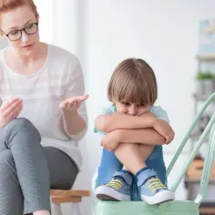 Kobieta w okularach rozmawia z chłopcem, który skulony siedzi na krześle. Nauczyciel wspomagający wspiera dzieci z trudnościami.