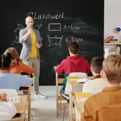 Nauczyciel, jak przewodniczący rady rodziców, tłumaczy zadanie z matematyki. Uczniowie uważnie słuchają, jeden z nich podnosi rękę.
