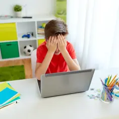 Chłopiec zamyka twarz w dłoniach, martwiąc się, czy można nie dostać się do żadnej szkoły. Na biurku laptop, zeszyty i lampka.