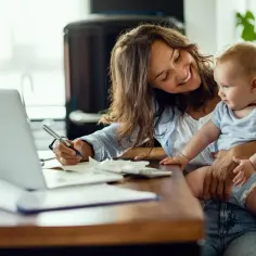 Uśmiechnięta mama z dzieckiem na rękach wypełnia dokumenty. Może to być wniosek o subwencję na dziecko z orzeczeniem o potrzebie kształcenia specjalnego.