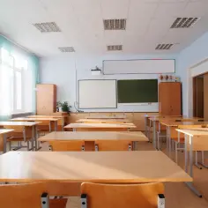 Pusta sala lekcyjna z rzędami drewnianych ławek i krzeseł. Na ścianie tablica, projektor i szafki. To typowa jednostka budżetowa, gotowa na przyjęcie uczniów.