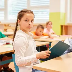 Dziewczynka z warkoczem czyta książkę, podczas gdy inni uczniowie siedzą przy ławkach. To część programu nauczania edukacja zdrowotna.