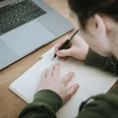 Uczeń pisze w zeszycie, obok leży laptop. Nauczyciel przygotowuje opinię o uczniu.