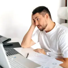 Mężczyzna z ręką na czole przy biurku z laptopami i dokumentami, może to być jego pierwszy staż pracy na umowie zlecenie.
