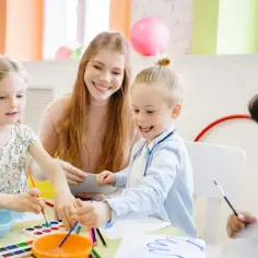 Uśmiechnięta pani pomaga dzieciom malować. To świetna zabawa, która może pomóc zdobyć za co punkty do przedszkola.