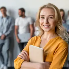Uśmiechnięta nauczycielka z książką, gotowa do rozwoju zawodowego zgodnie z rozporządzeniem. W tle grupa osób.