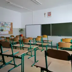 Pusta sala lekcyjna z rzędami krzeseł i biurek, tablicą i projektorem.