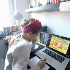 Dziewczynka z opaską na głowie uczy się zdalnie, oglądając bajkę na laptopie. W tle półka z książkami i kalendarz.