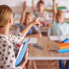Nauczycielka w serduszka wskazuje na ucznia. Dzieci podnoszą ręce. Nawet w wakacje zwolnienie lekarskie nauczyciela może być potrzebne.