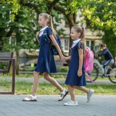 Dwie dziewczynki idą chodnikiem, trzymając się za ręce. Mają na sobie granatowe sukienki i plecaki. W tle chłopiec jedzie na rowerze. To obrazek, który przypomina kolejność przedmiotów na świadectwie.