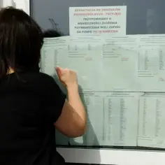 Kobieta sprawdza listę kandydatów do szkoły średniej. Ogłoszenie przypomina o potwierdzeniu woli zapisu.