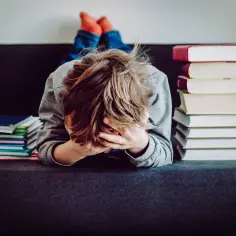 Chłopiec z rękami na twarzy, przytłoczony stertą książek. Może to znak, że potrzebne jest orzeczenie z poradni psychologiczno-pedagogicznej.