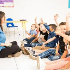 Nauczycielka siedzi na podłodze, a dzieci z podniesionymi rękami czekają na swoją kolej. Trwa rekrutacja uzupełniająca do przedszkola.