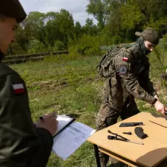 Kandydaci do szkoły mundurowej ćwiczą rozkładanie broni. Na stole leżą części karabinu, a jeden z żołnierzy sprawdza listę.