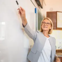 Nauczycielka dyplomowana z markerem przy tablicy, jak w przykładowej ocenie pracy. Uśmiechnięta, w okularach, w szarej marynarce.
