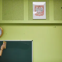 Klasa z zegarem, godłem Polski i krzyżem. Ręka zmywa napis z tablicy, jakby to był wzór rezygnacji ze szkoły podstawowej.