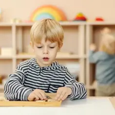 Chłopiec z blond włosami skupiony na zadaniu z materiałami do nauki metodą Montessori. W tle inne dziecko i półki z zabawkami.