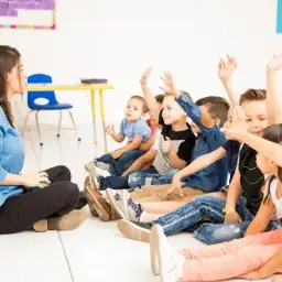 Dzieci na lekcji z podniesionymi rękami. Nauczycielka siedzi na podłodze. Frekwencja a ocena z zachowania - czy obecność wpływa na ocenę?