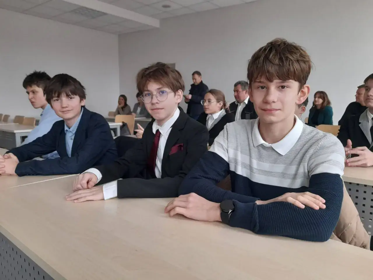 Trzech chłopców siedzi w ławkach szkolnych, skupionych na lekcji. Ich uśmiechy sugerują, że obowiązek nauki jest dla nich przyjemnością.