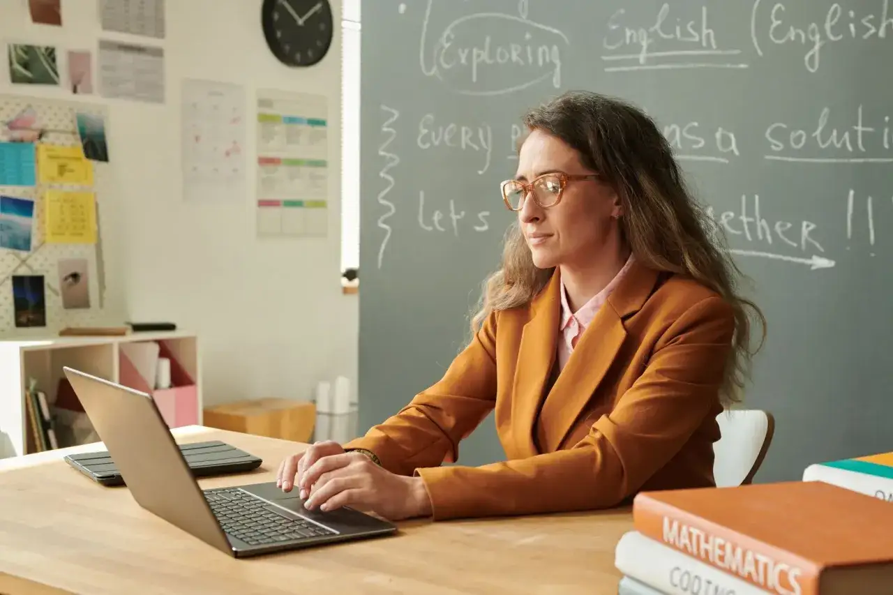 Nauczycielka w okularach pracuje przy laptopie. Na tablicy napisy po angielsku, a na biurku książki. Widać, że to jej wysługa lat nauczyciela.