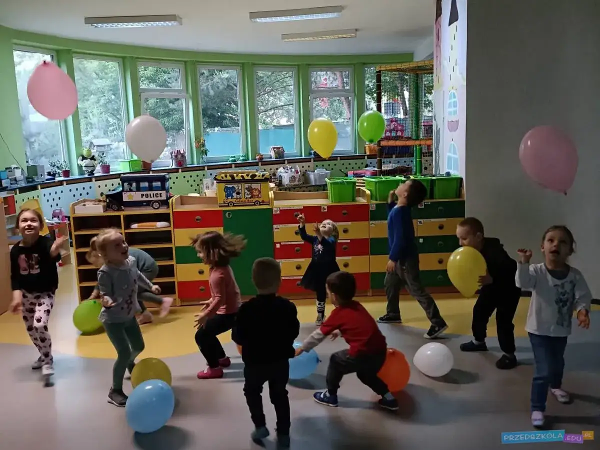 Dzieci bawią się balonami w sali, jak w przedszkolu. Radość i ruch w punkcie przedszkolnym.