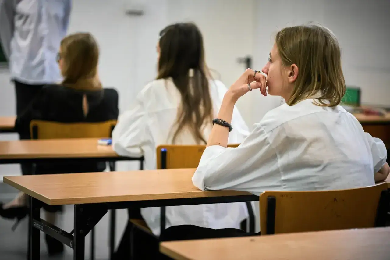 Dziewczyna zamyślona podczas egzaminu klasyfikacyjnego. W tle inni uczniowie siedzą przy ławkach.