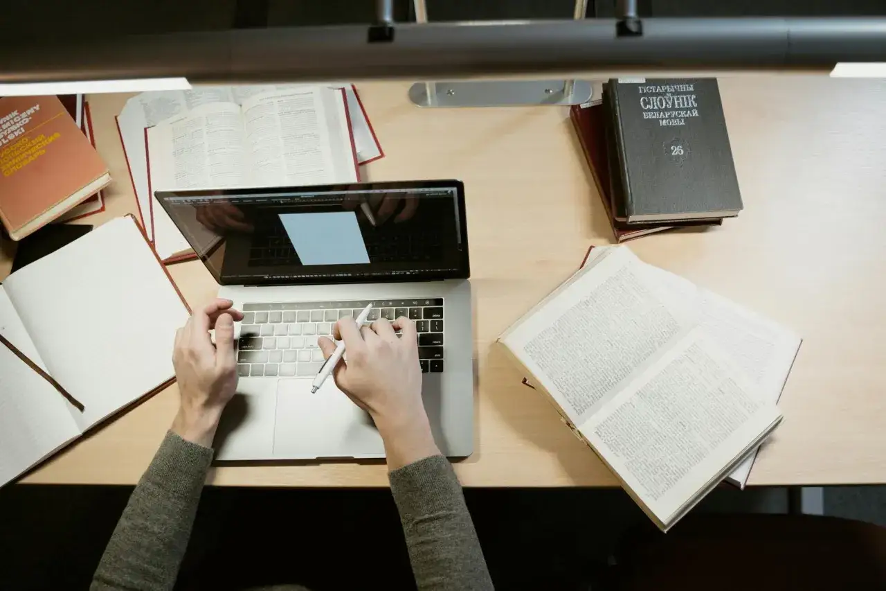 Ręce piszą na laptopie, otoczone książkami i notatnikiem. Nauka i praca, ale czy to nie jest odkładanie na później?