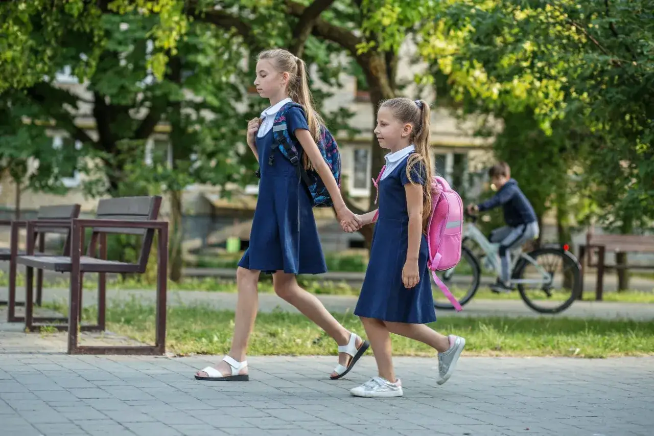 Dwie dziewczynki w granatowych sukienkach i z plecakami idą chodnikiem. Starsza trzyma za rękę młodszą. W tle chłopiec na rowerze. Kolejność przedmiotów na świadectwie jest ważna.