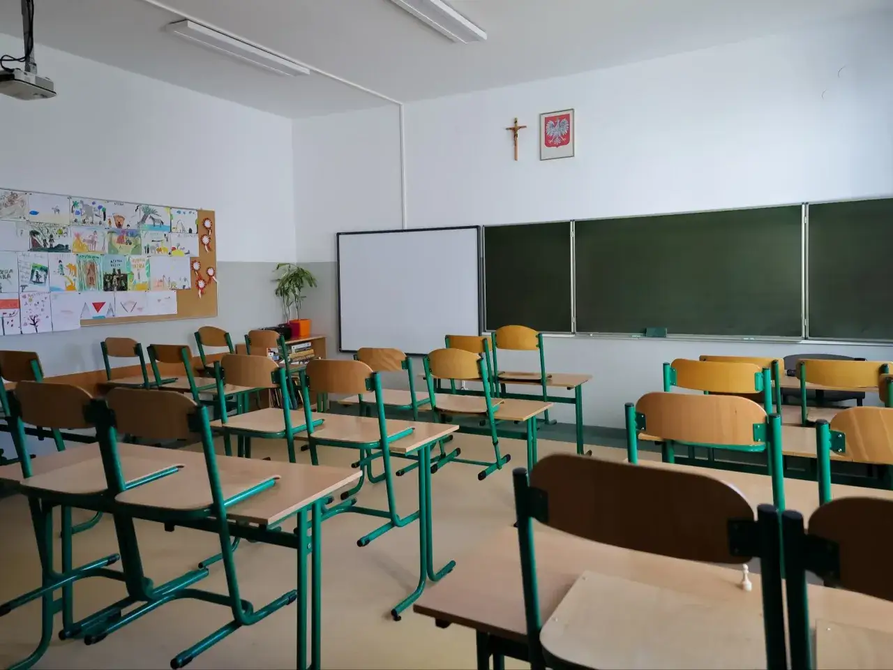 Puste ławki w sali lekcyjnej, gotowe na pismo do rodziców w sprawie realizacji obowiązku szkolnego. Na ścianie rysunki dzieci i godło Polski.