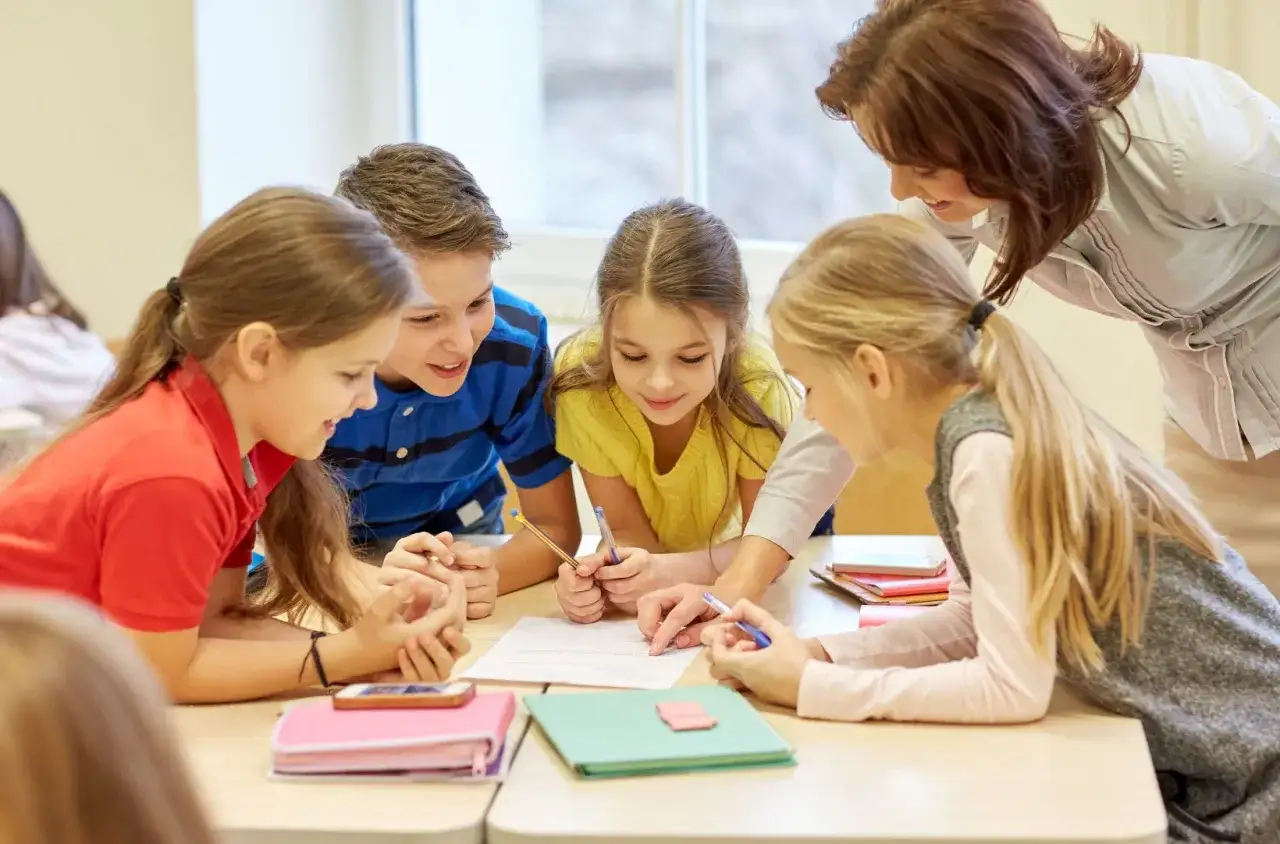 Nauczycielka pomaga uczniom z dysleksją, omawiając zadania. Dzieci skupione na pracy, widoczne objawy dysleksji, jak trudności z czytaniem, mogą być wspierane.