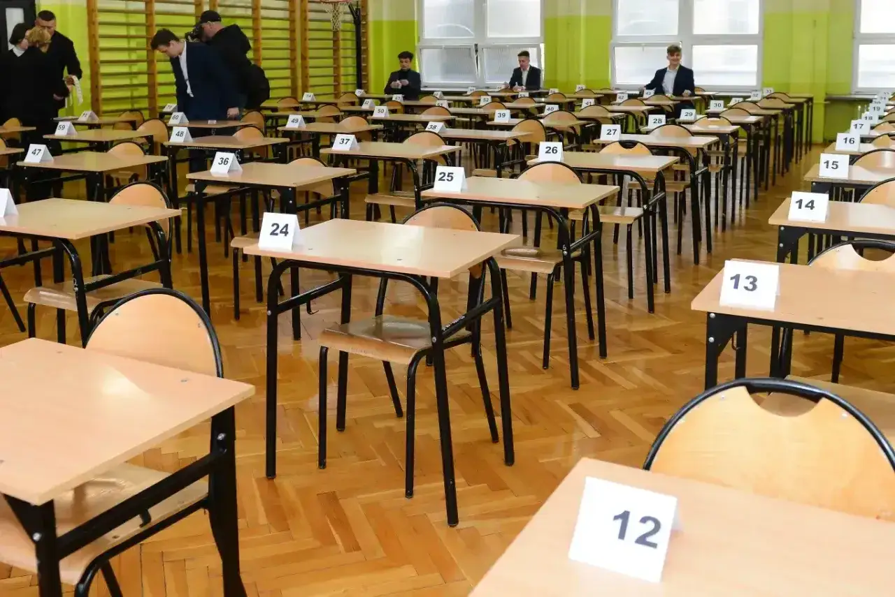 Puste ławki w sali gimnastycznej, przygotowane do egzaminu ósmoklasisty. Numery na stolikach sugerują wydłużony czas na egzaminie.
