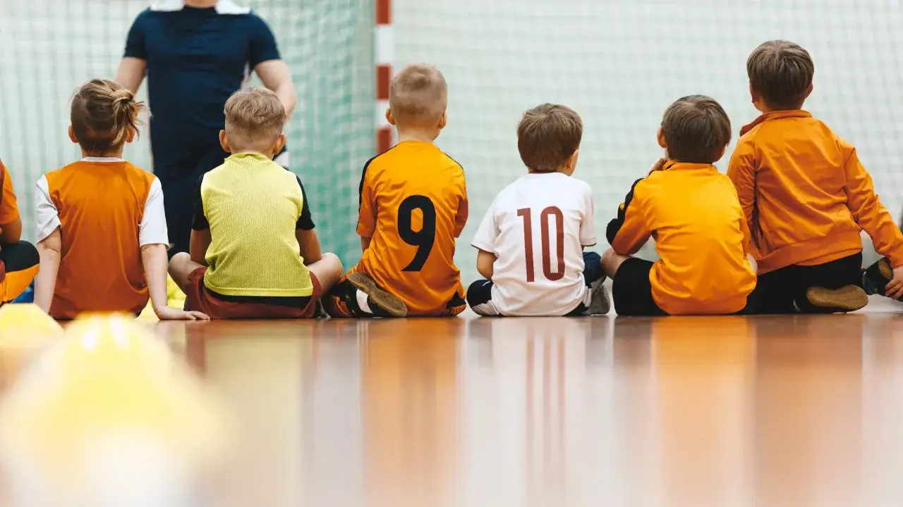Dzieci w sportowych strojach siedzą na podłodze sali gimnastycznej. To klasa sportowa w podstawówce, gotowa do treningu.