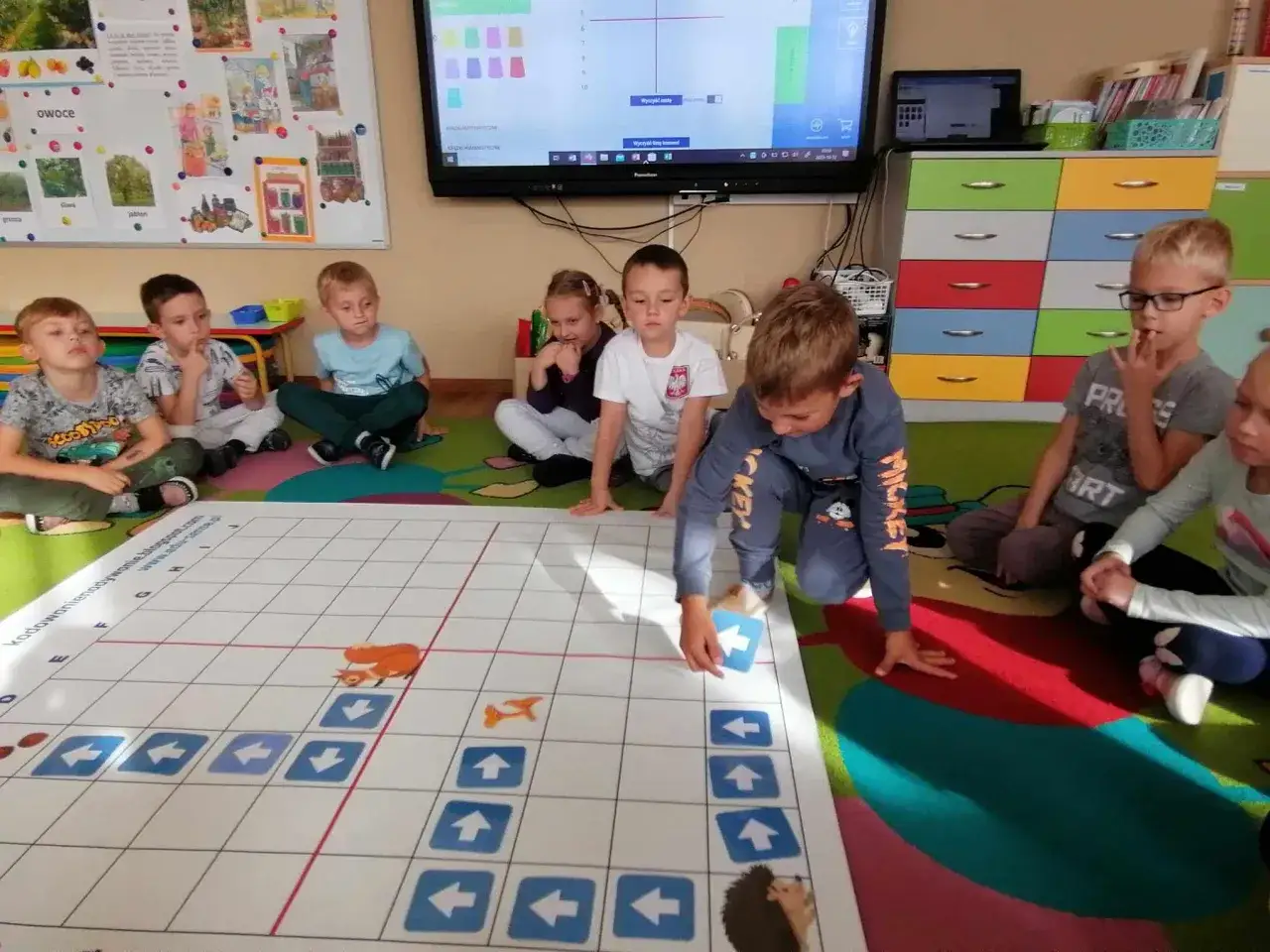 Dzieci uczą się kodowania na dywanie, rozwiązując zadanie z liskiem. W przedszkolu bezpłatnych godzin jest wiele, a zabawy edukacyjne to świetny sposób na naukę.