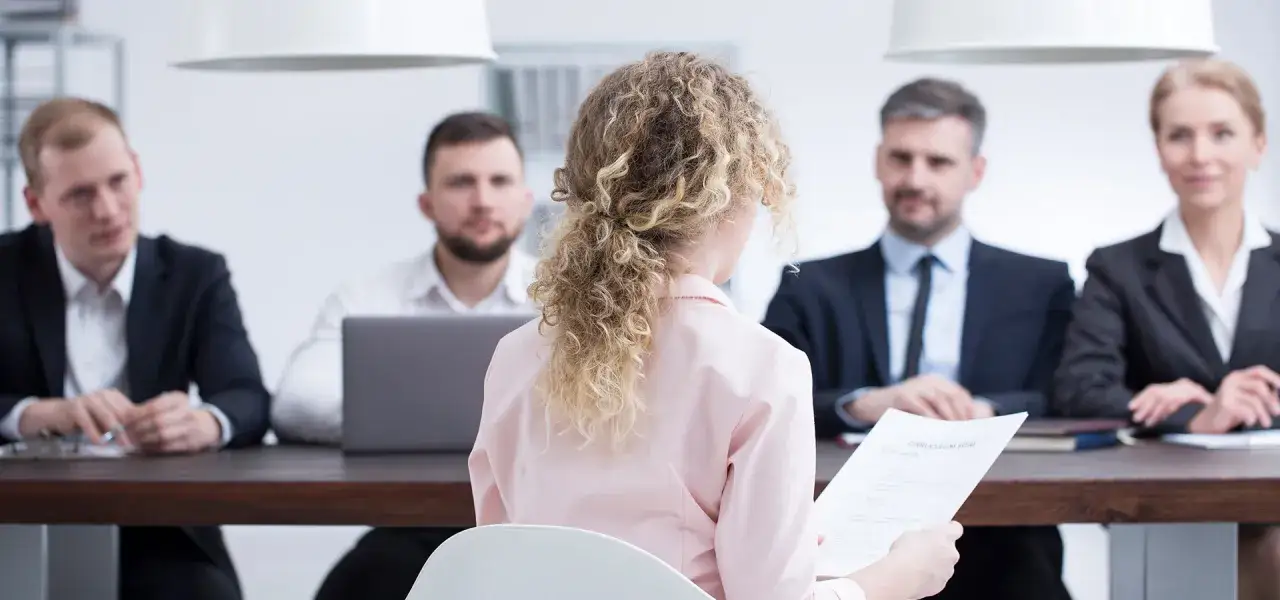 Kobieta z kręconymi włosami trzyma CV przed komisją. Czy ktoś oblał egzamin na dyplomowanego?