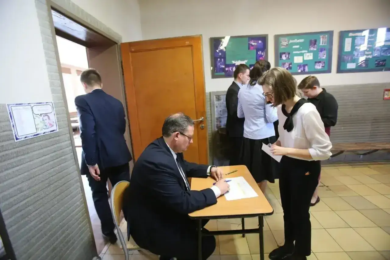 Uczeń w garniturze wchodzi do sali, gdzie egzaminator sprawdza dokumenty. To ważny moment, jak przygotować się do matury ustnej z polskiego.
