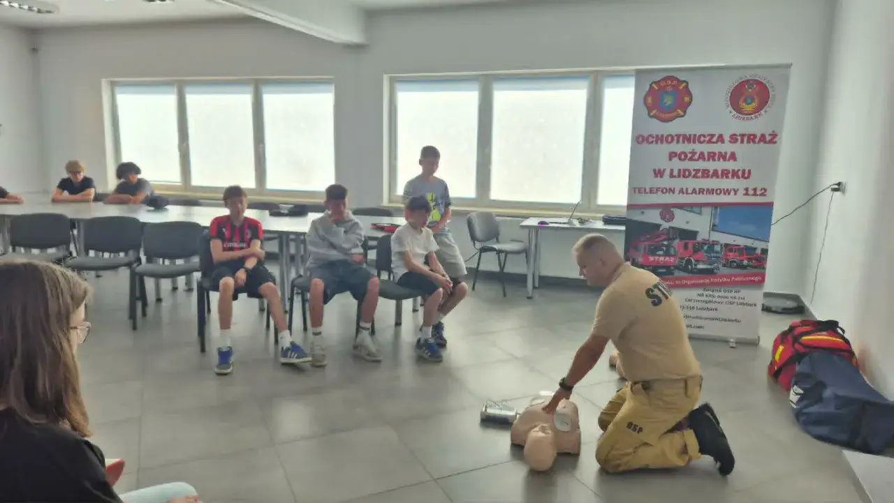 Próbna ewakuacja w OSP Lidzbark. Strażak demonstruje resuscytację na fantomie, młodzież obserwuje.